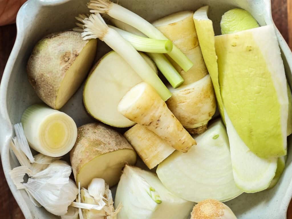 A bowl filled with various chopped vegetables, including potatoes, leeks, carrot, radish peel, and garlic cloves, all arranged closely together.